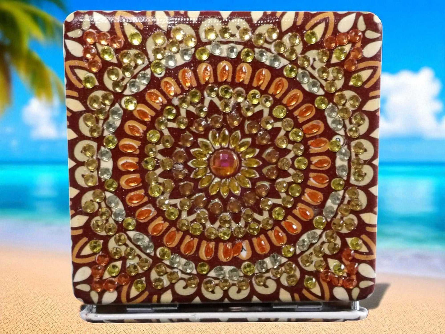 Brown square pocket mirror with mandala design and decorative gems on a beach background
