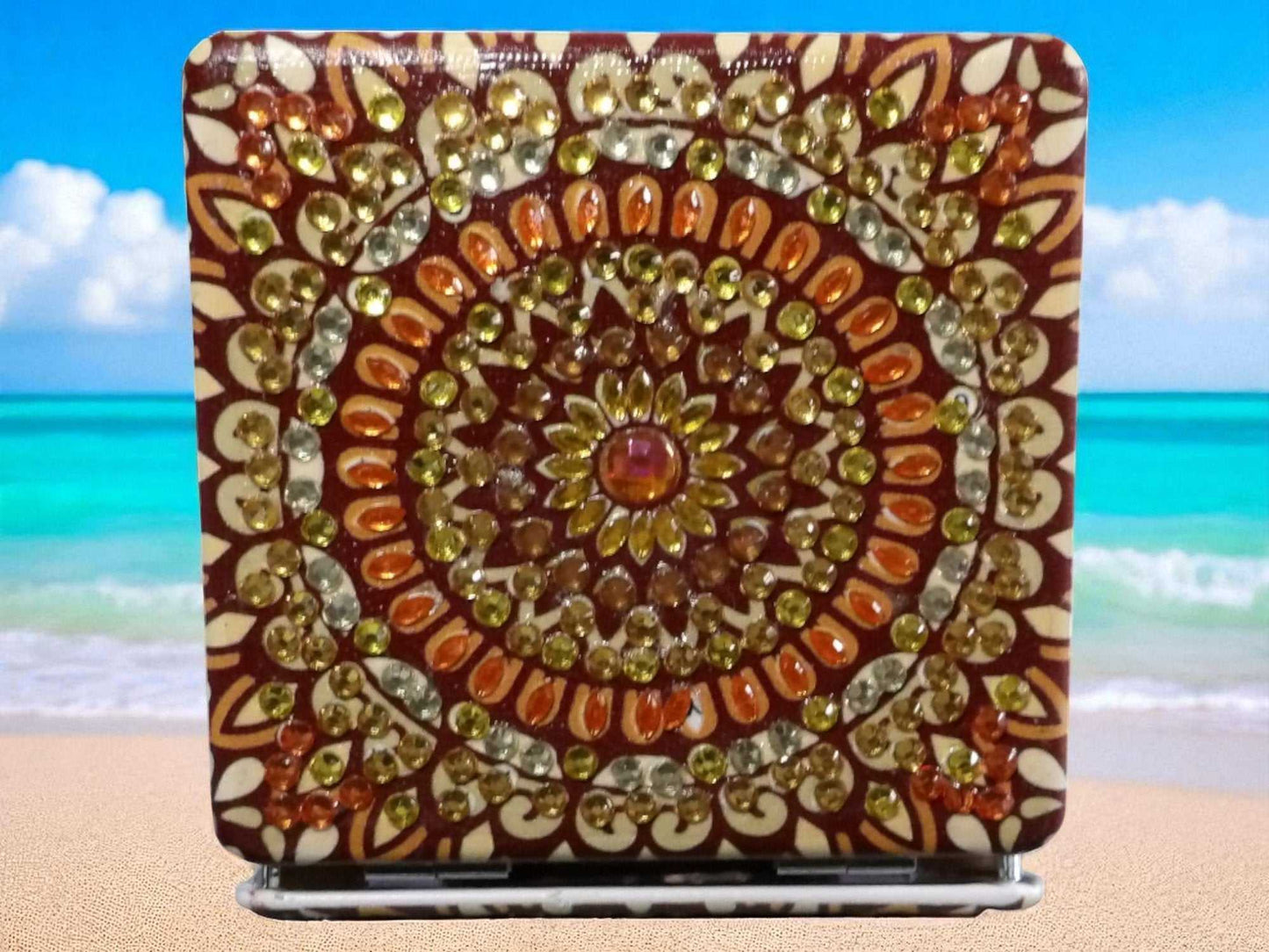 Brown square pocket mirror with mandala design and decorative gems on beach background