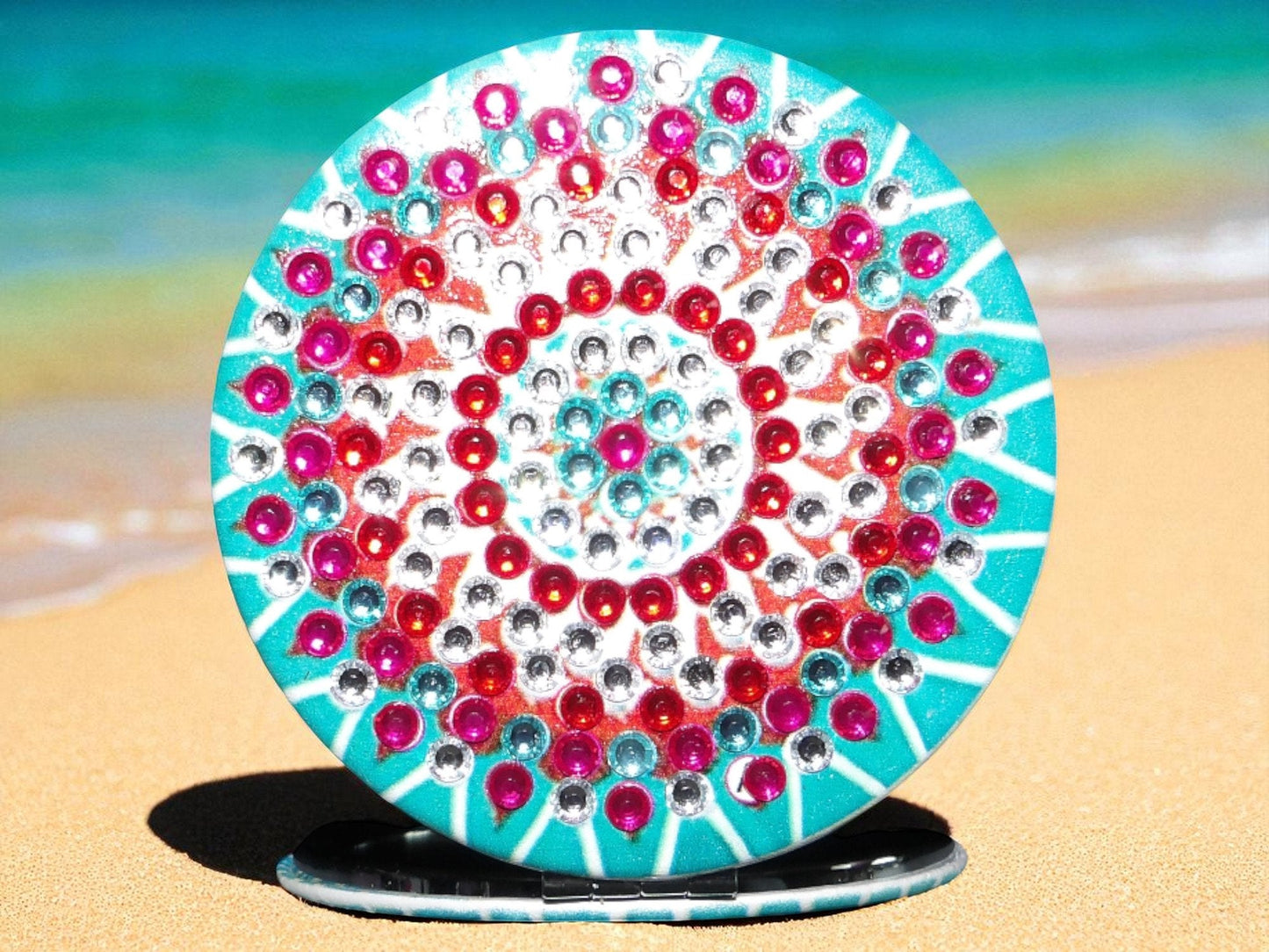 Blue and red round pocket mirror with mandala design and magnification on one side on beach sand background