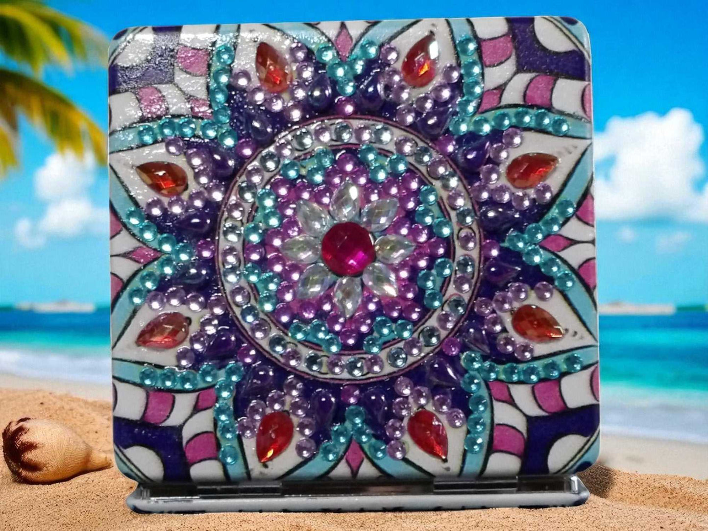 Purple square pocket mirror with colorful mandala design and gemstones on sandy beach background