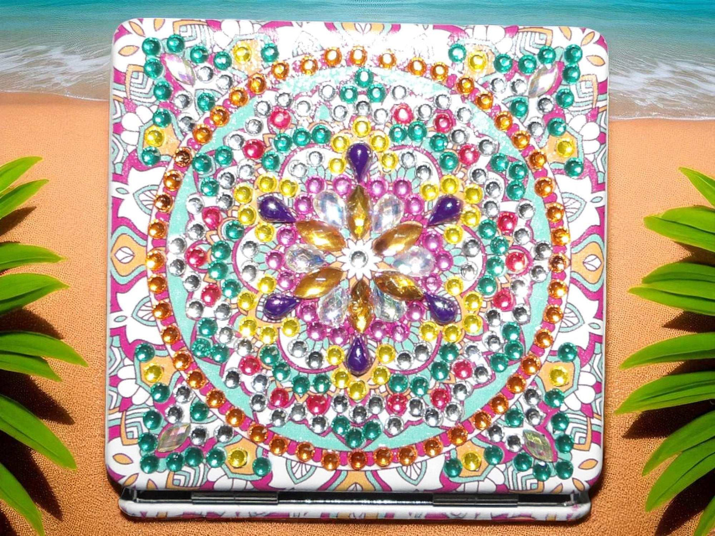 Greenish square pocket mirror with colorful mandala design and rhinestone embellishments on a beach background