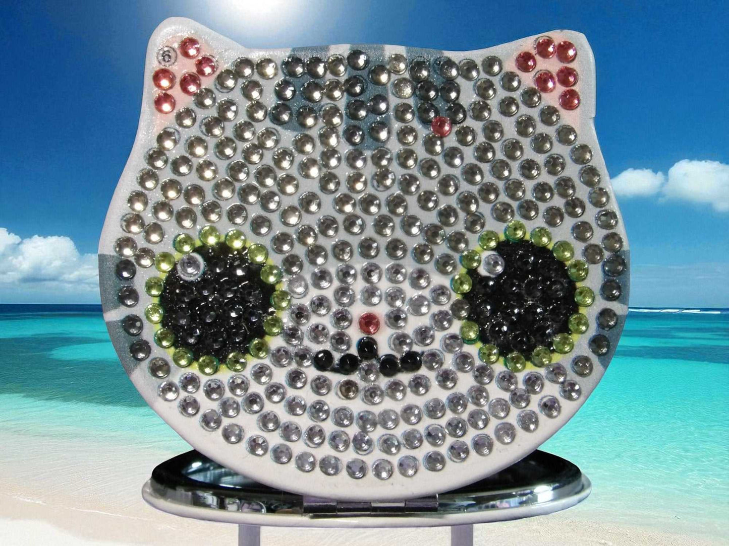 Silver cat face pocket mirror with rhinestone design and magnification feature on tropical beach background