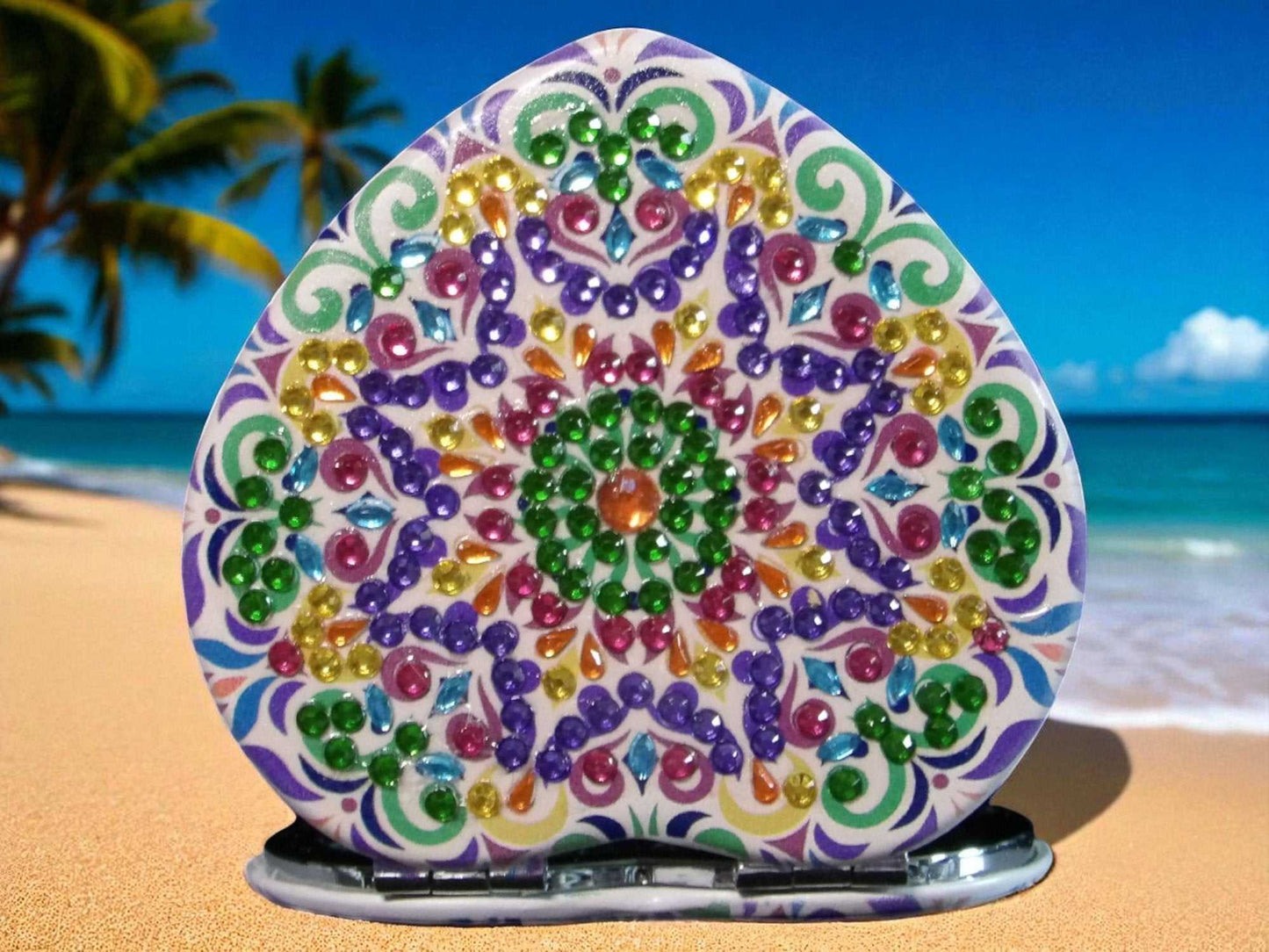 Purple and green heart shaped pocket mirror with colorful mandala design and magnification feature on tropical beach background
