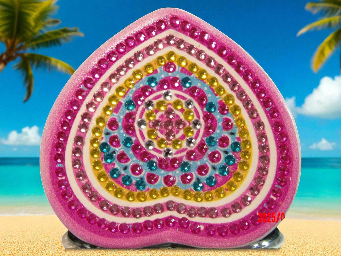 Pink and yellow heart shaped pocket mirror with mandala design and colorful rhinestones on beach background