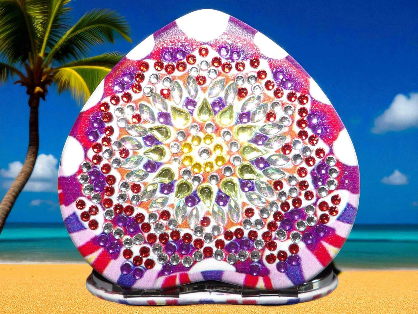Pink and purple heart shape pocket mirror with mandala design and decorative gemstones on beach background