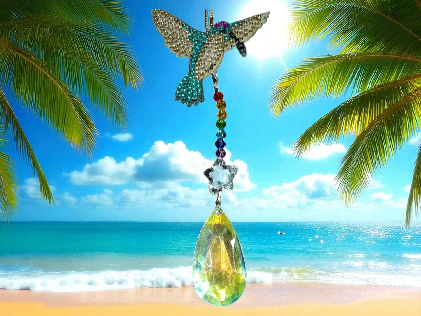 Blue & white hummingbird sun catcher with crystal drops hanging against a tropical beach background