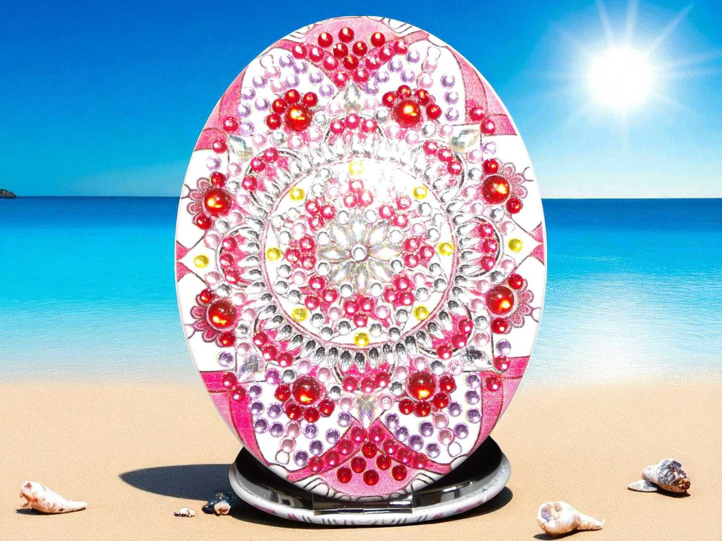 Red oblong pocket mirror with mandala design and decorative red and pink beads on beach background