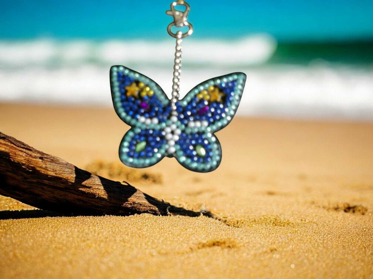 Dark and light blue butterfly keychain hanging on a beach driftwood with ocean waves in the background