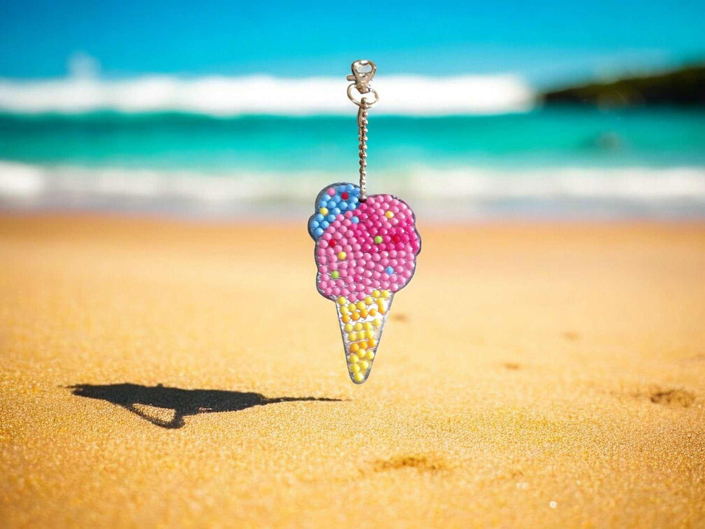 Colorful beaded ice cream cone keychain hanging above sandy beach with ocean background