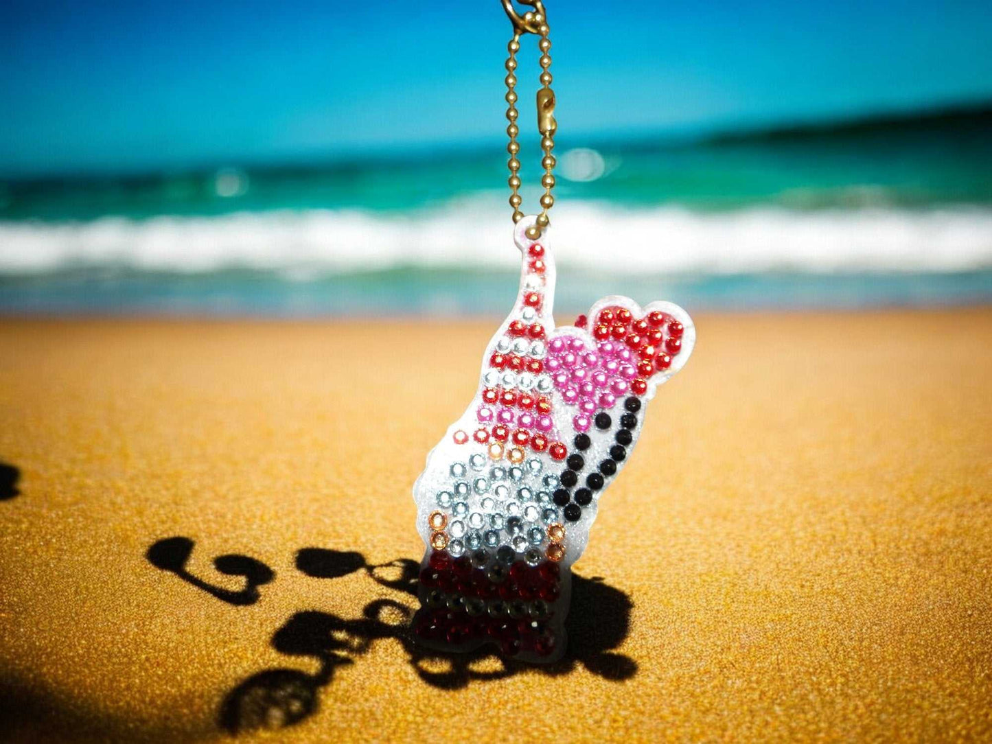 Gnome wHeart Balloons keychain with colorful rhinestones hanging on a gold chain at the beach sand with ocean background
