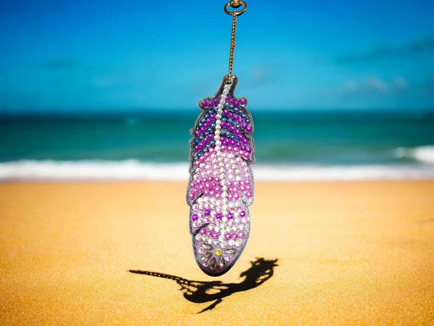 Pink and purple feather keychain with beads hanging outdoors on a sunny beach background