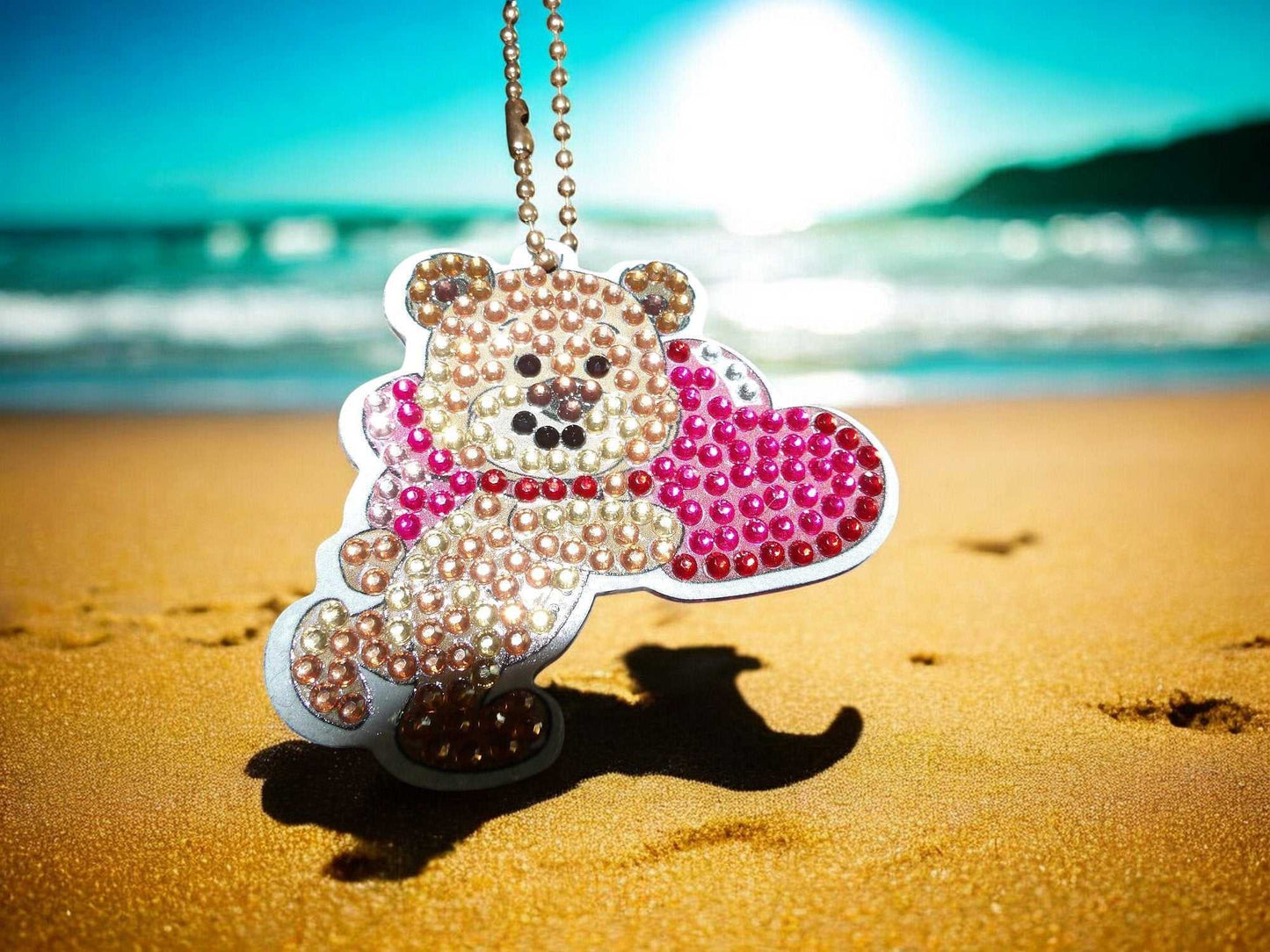 Running teddy bear keychain holding a pink heart with sparkling beads on a beach sand background