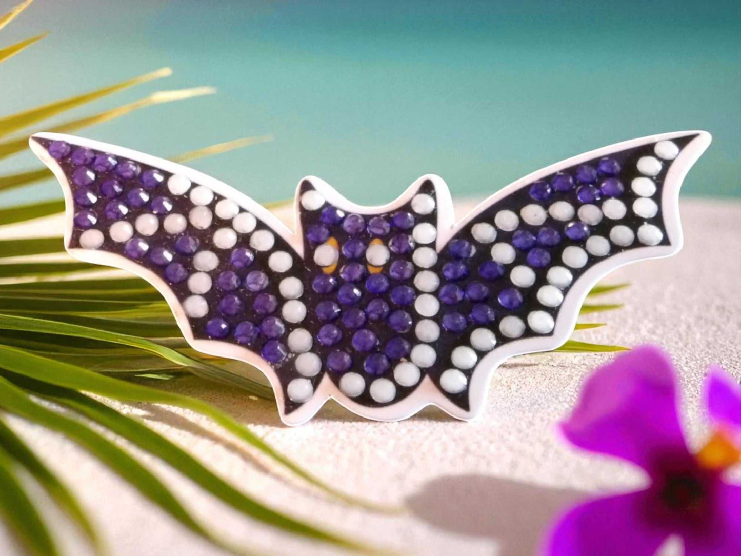 Glow in the dark flying bat magnet with purple and white beads on sandy surface