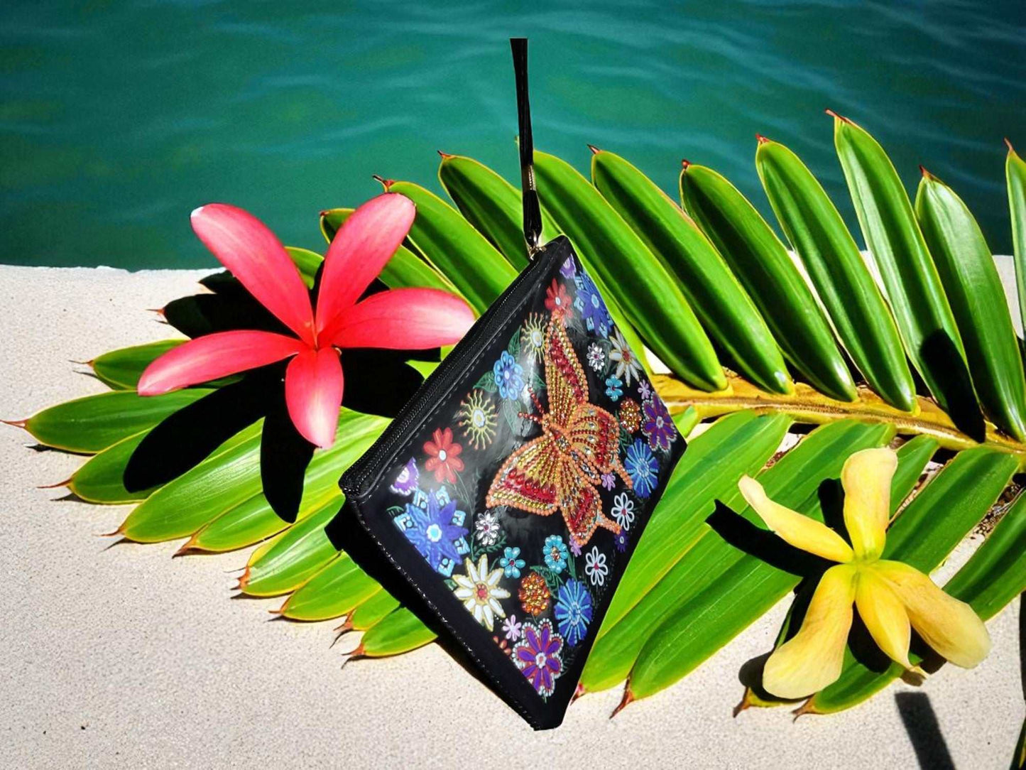 Golden Butterfly Large Hand Clutch with colorful butterfly and floral design on black background placed on tropical leaves with pink and yellow flowers