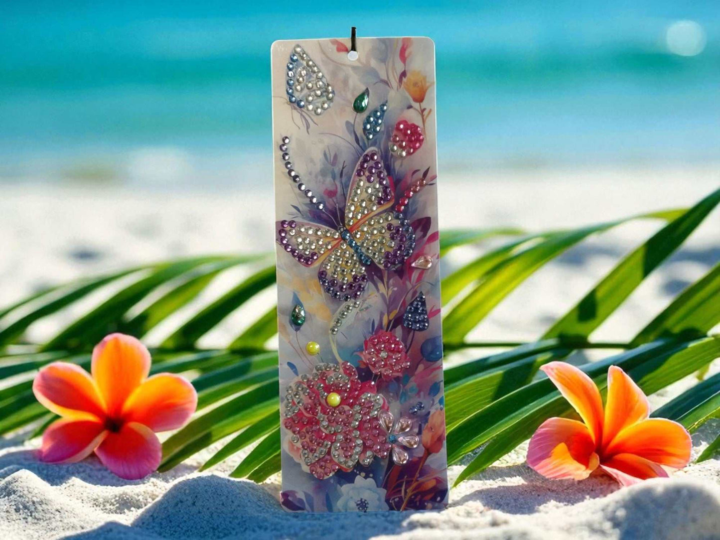 Purple and gold butterfly colorful bookmark with floral design and rhinestone accents on a tropical beach background
