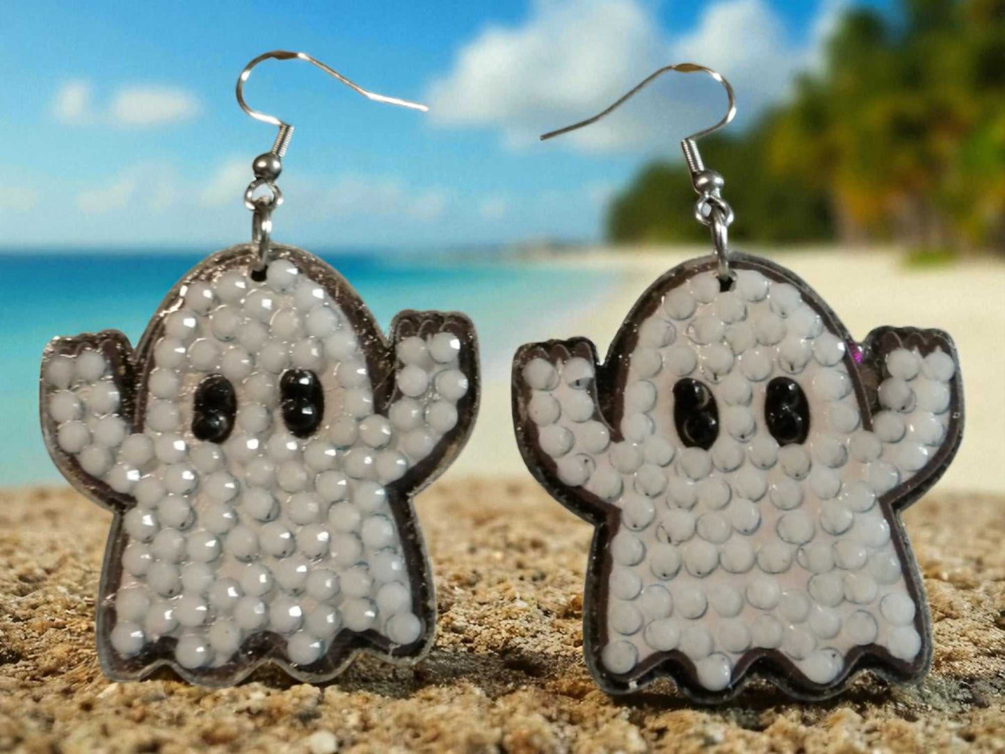 Glow in the dark ghost earrings with white beaded design and black eyes on sandy beach background