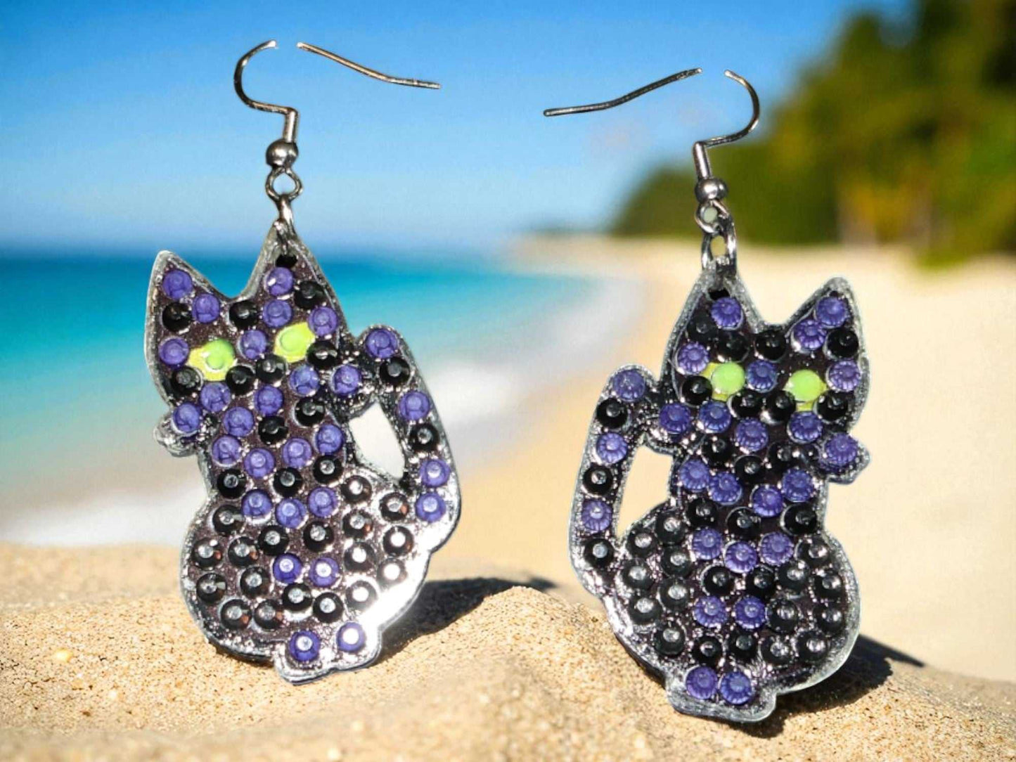 Glow in the dark black cat earrings with purple and green accents on a beach background