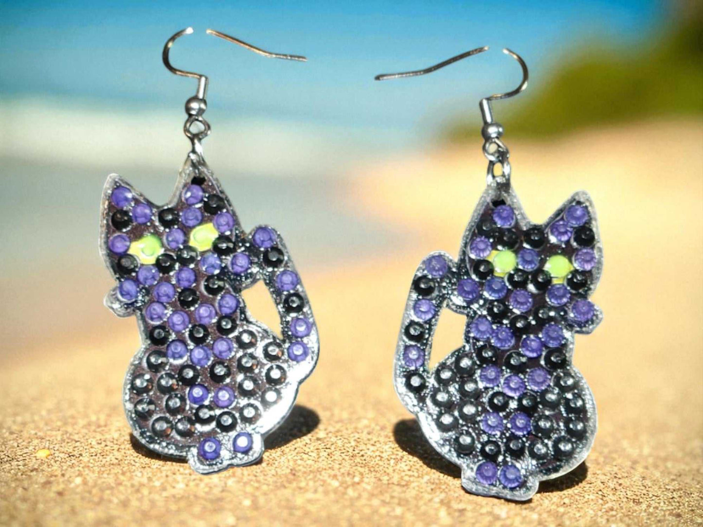 Glow in the dark black cat earrings with purple and green accents on a beach background