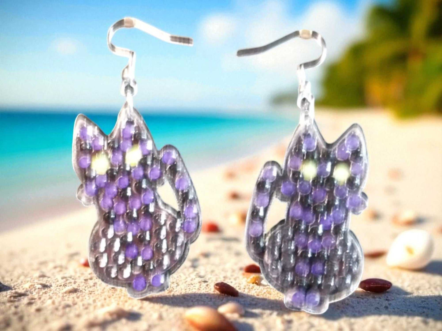 Glow in the dark black cat earrings with purple and green accents on a beach background