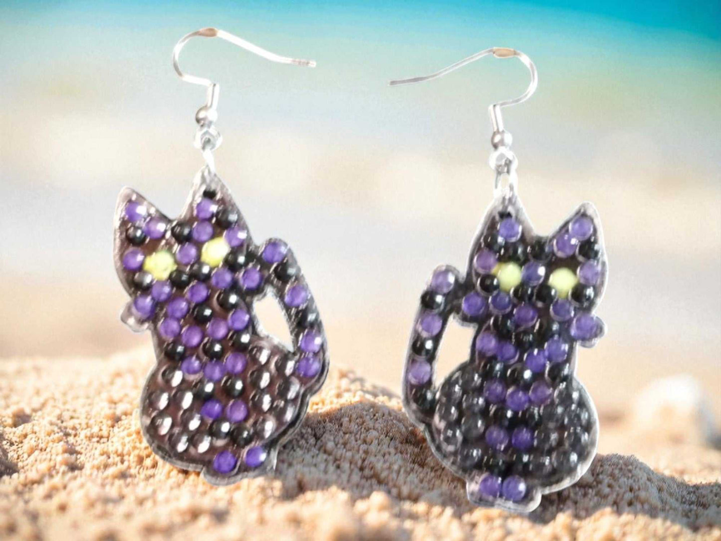 Glow in the dark black cat earrings with purple and green accents on a beach background