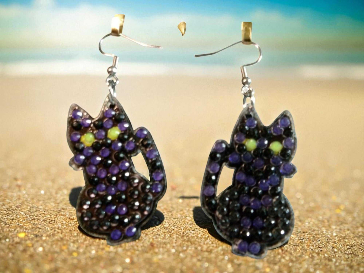 Glow in the dark black cat dangle earrings with purple and yellow accents on sandy beach background