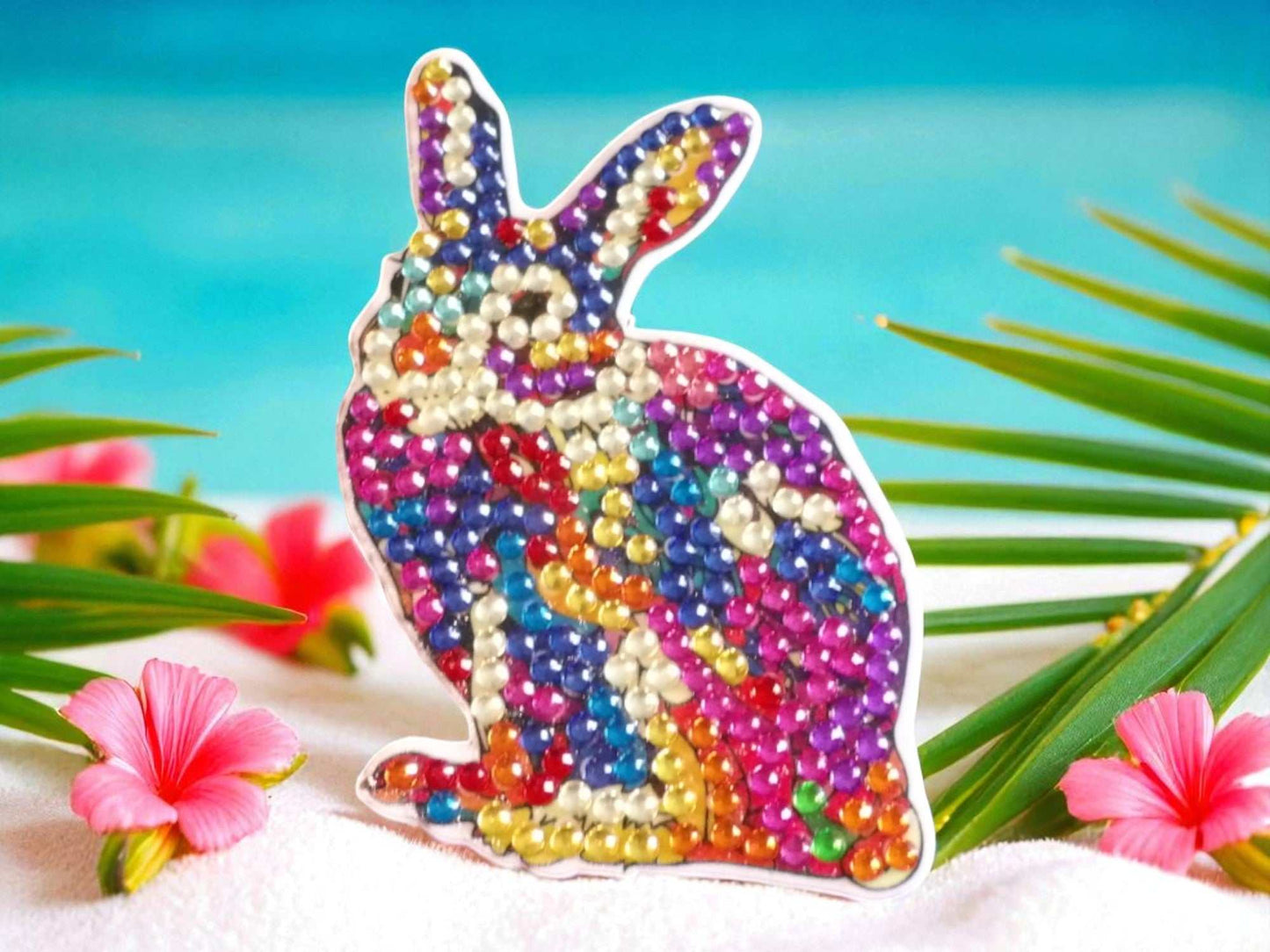 Colorful rabbit magnet decorated with vibrant rhinestones on a beach setting with flowers and palm leaves