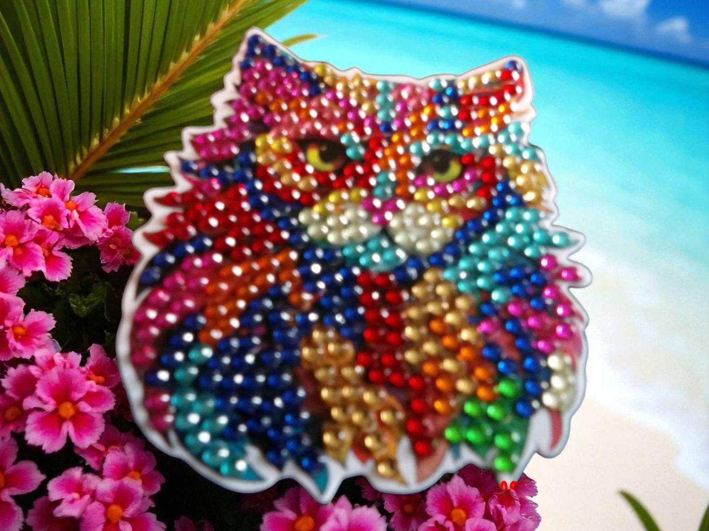 Colorful cat face magnet with vibrant beads near pink flowers and beach background