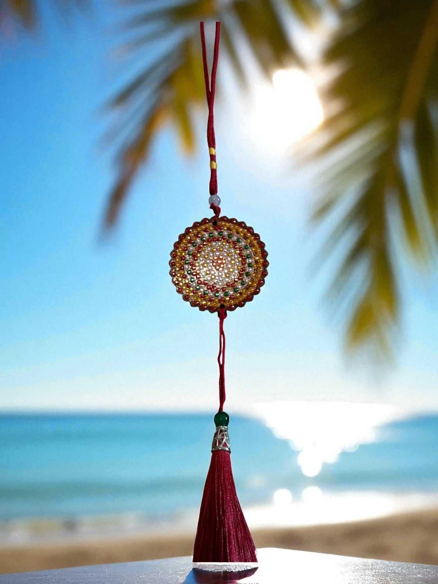 Deep red colorful design car ornament hanging with beaded circular pattern and tassel against beach background