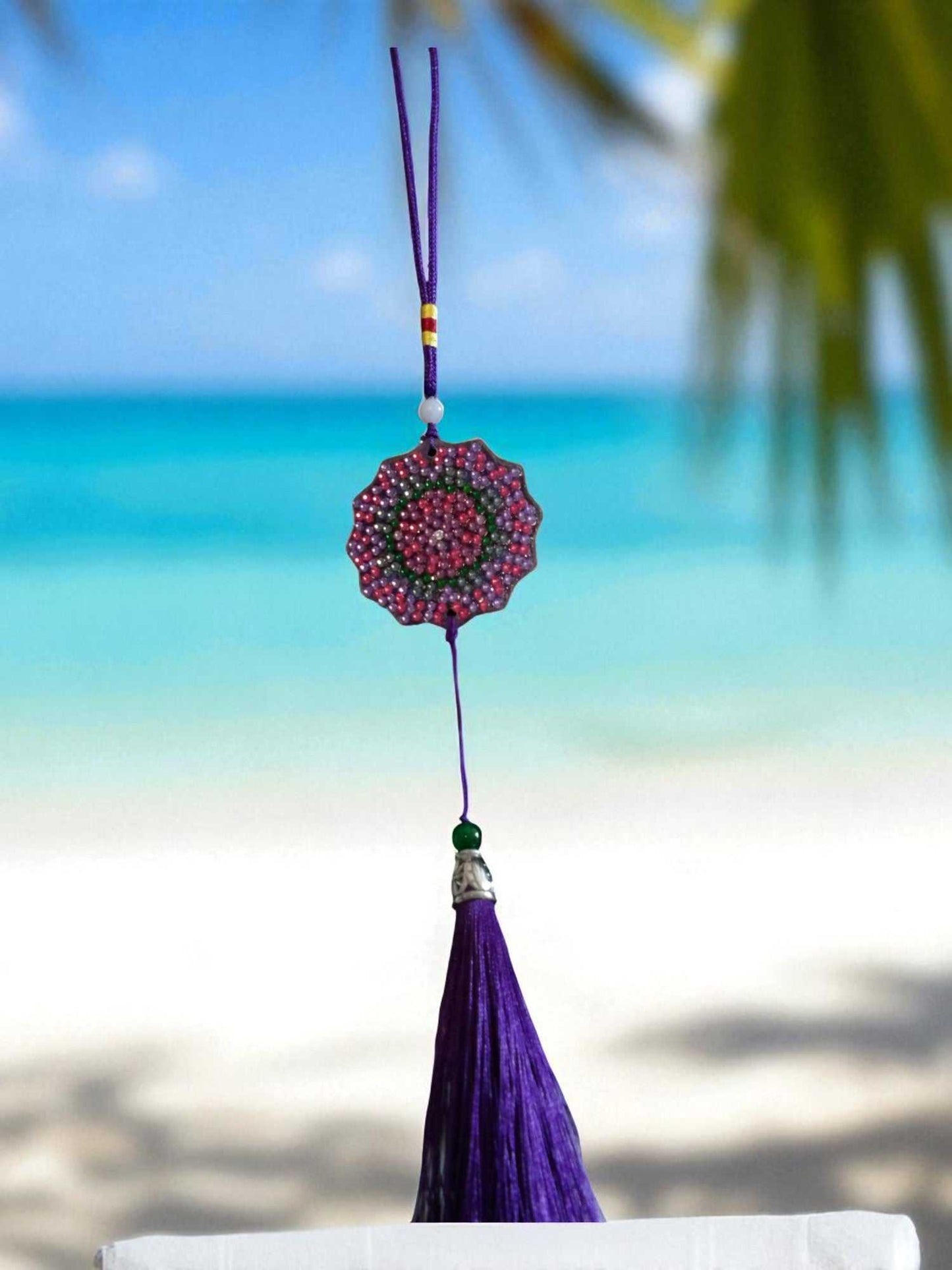 Deep blue colorful design car ornament with purple tassel hanging against a beach background