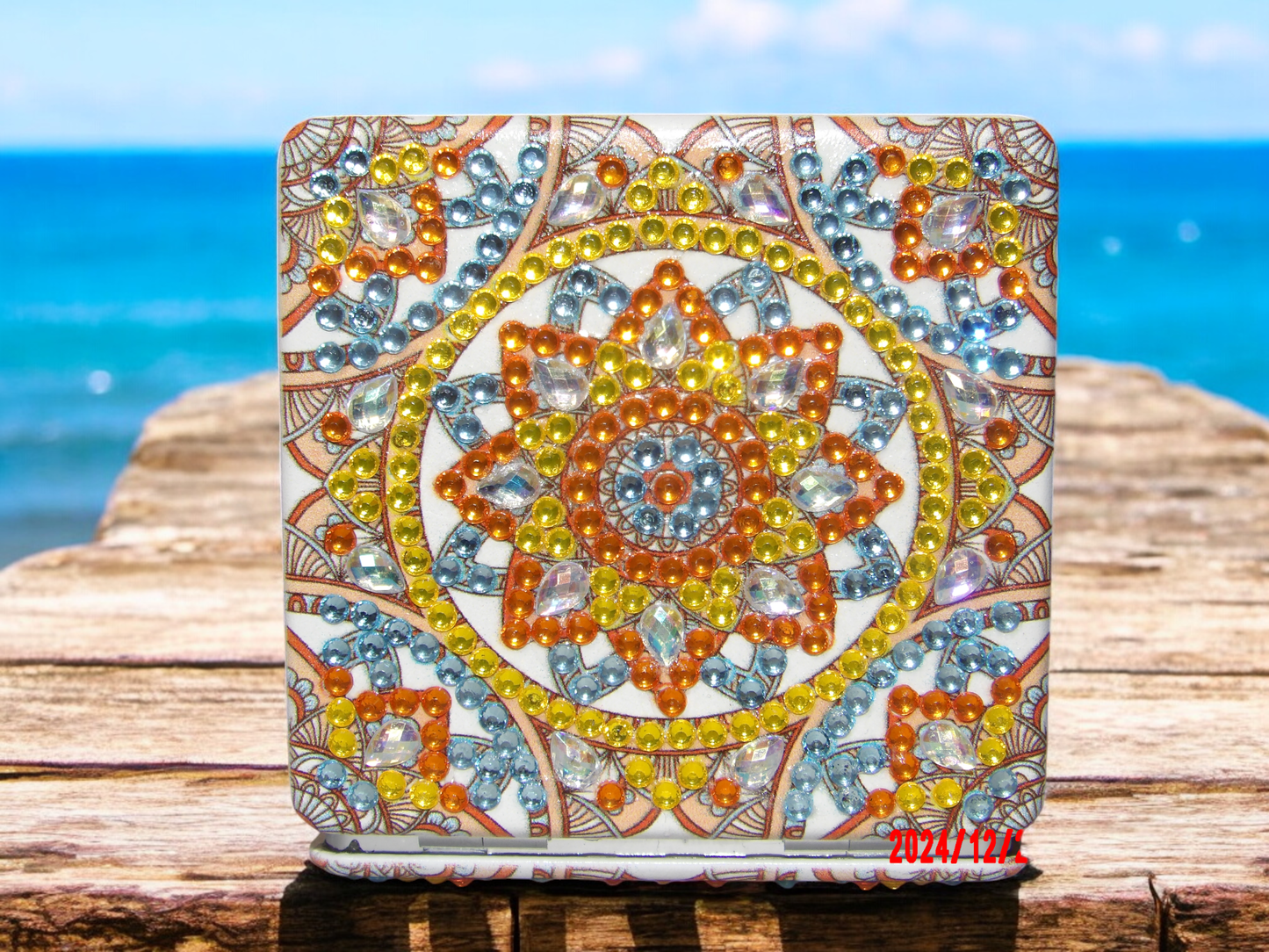 Compact golden mandala design pocket mirror with colorful gemstone embellishments on wooden surface by the sea