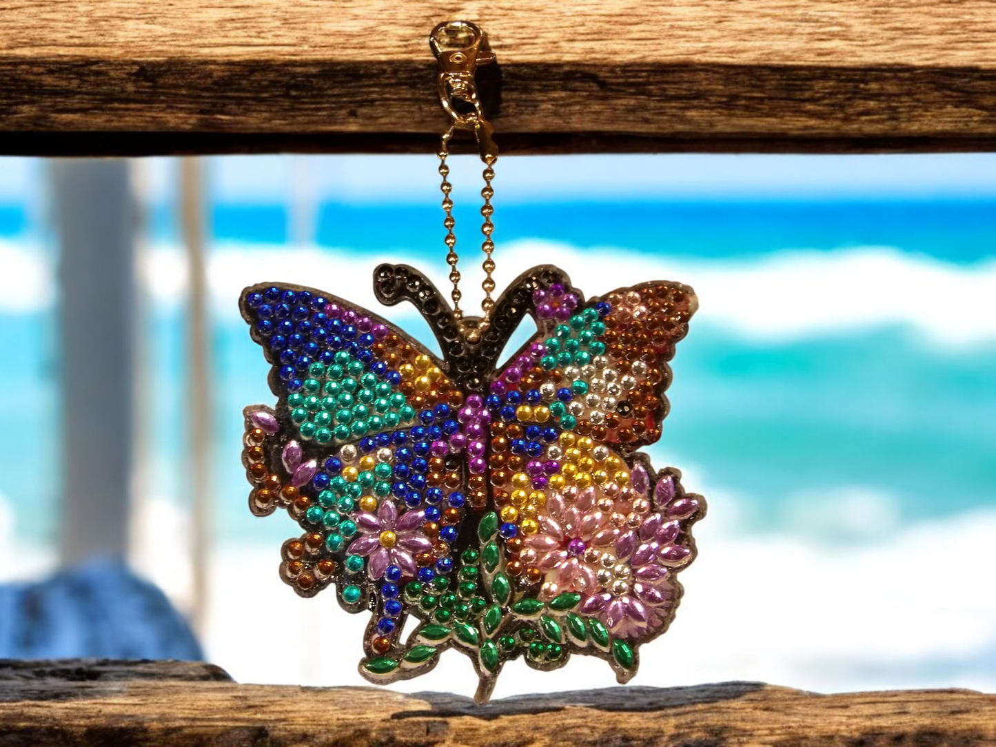 Large multi-color butterfly keychain with decorative beads hanging on a wooden frame against a blurred ocean background