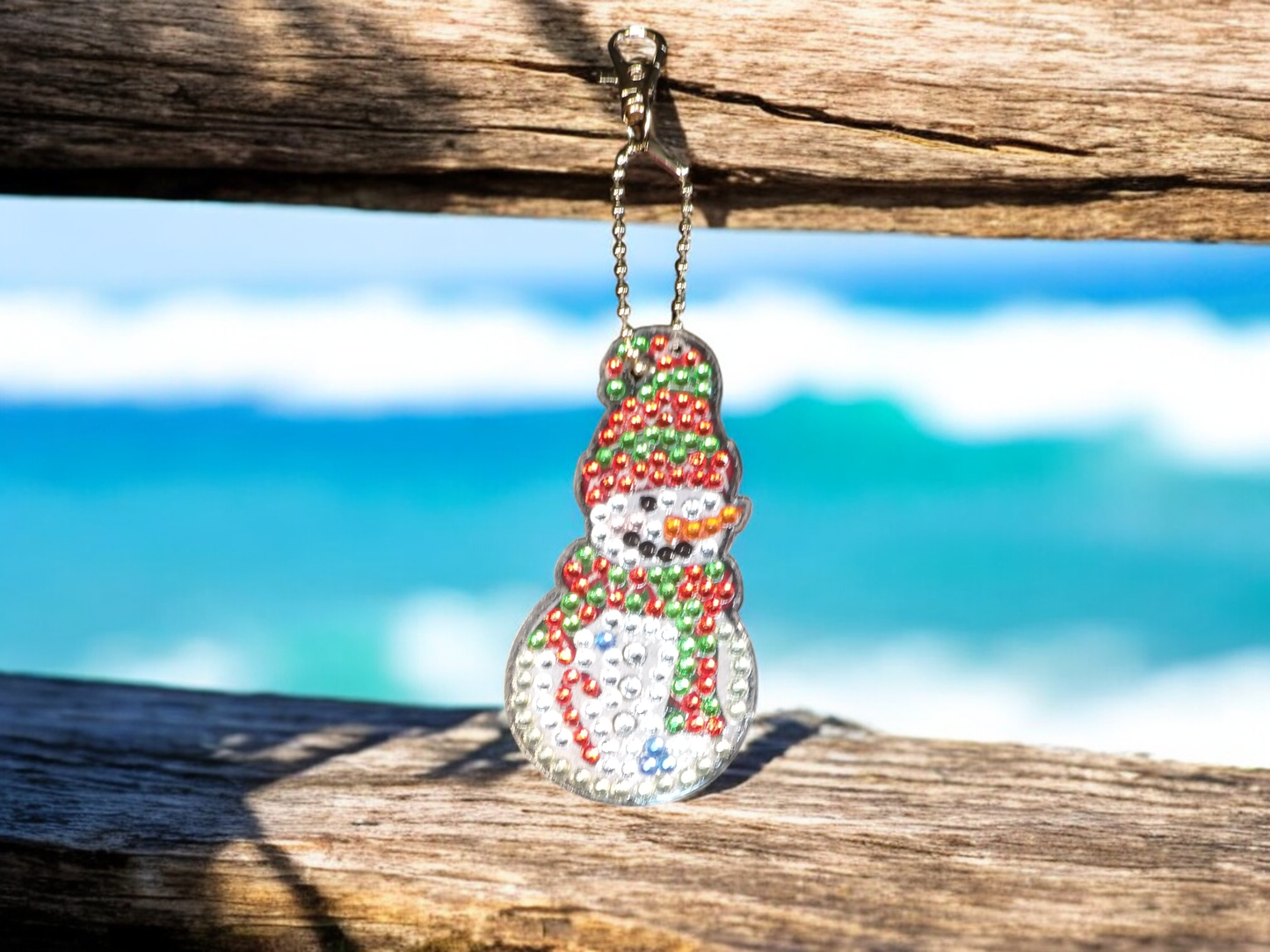 Red and green hat and scarf snowman keychain hanging on wooden surface with blurred ocean background