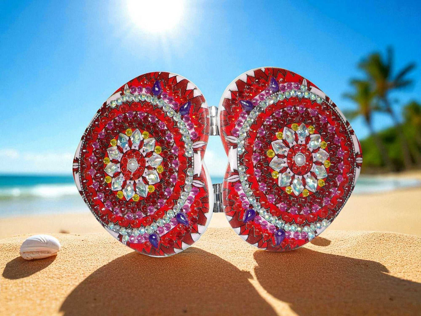 Red and pearl heart shape pocket mirror with mandala design and magnification on one side on sandy beach background
