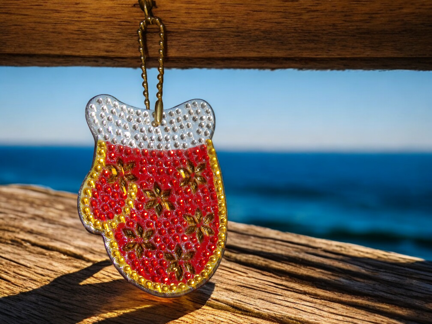 Red snow mitten keychain with decorative beads hanging on wooden surface with ocean background
