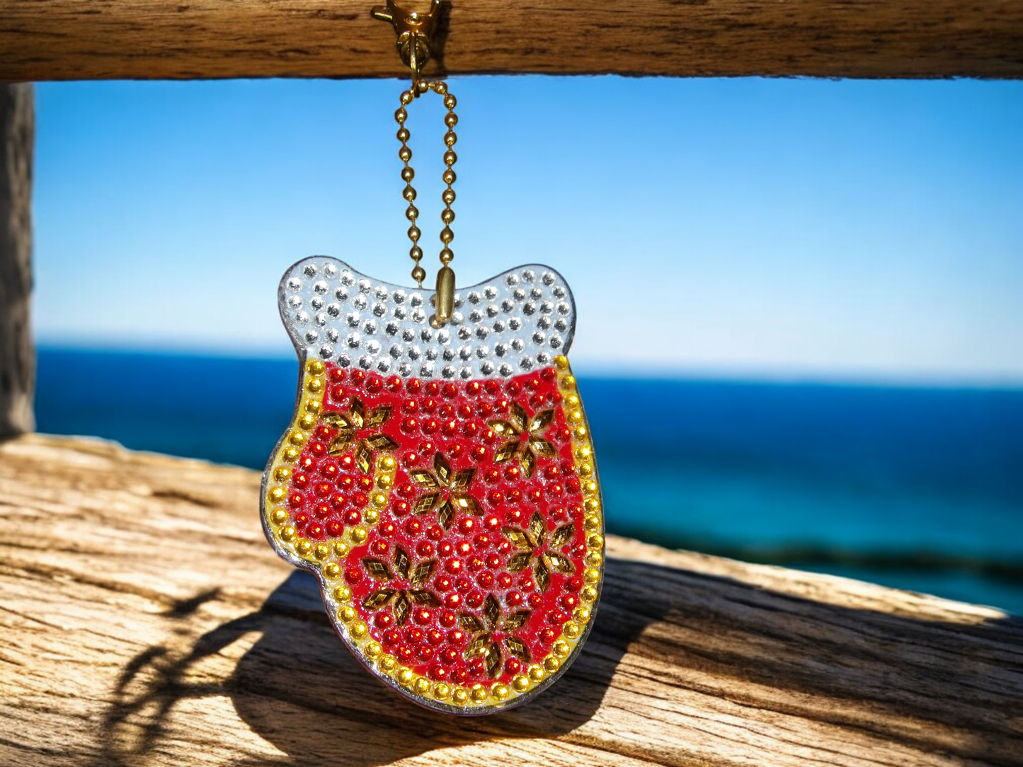 Red snow mitten keychain with gold and silver bead embellishments hanging on wooden railing by the ocean