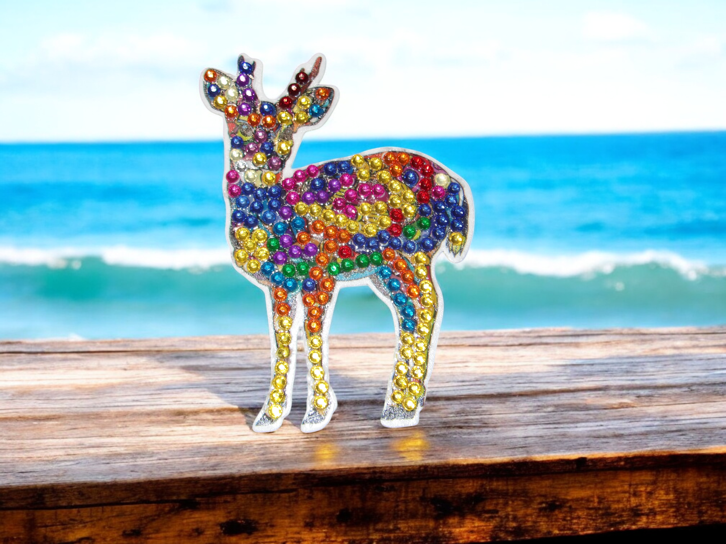 Young deer magnet decorated with colorful rhinestones on wooden surface by ocean background