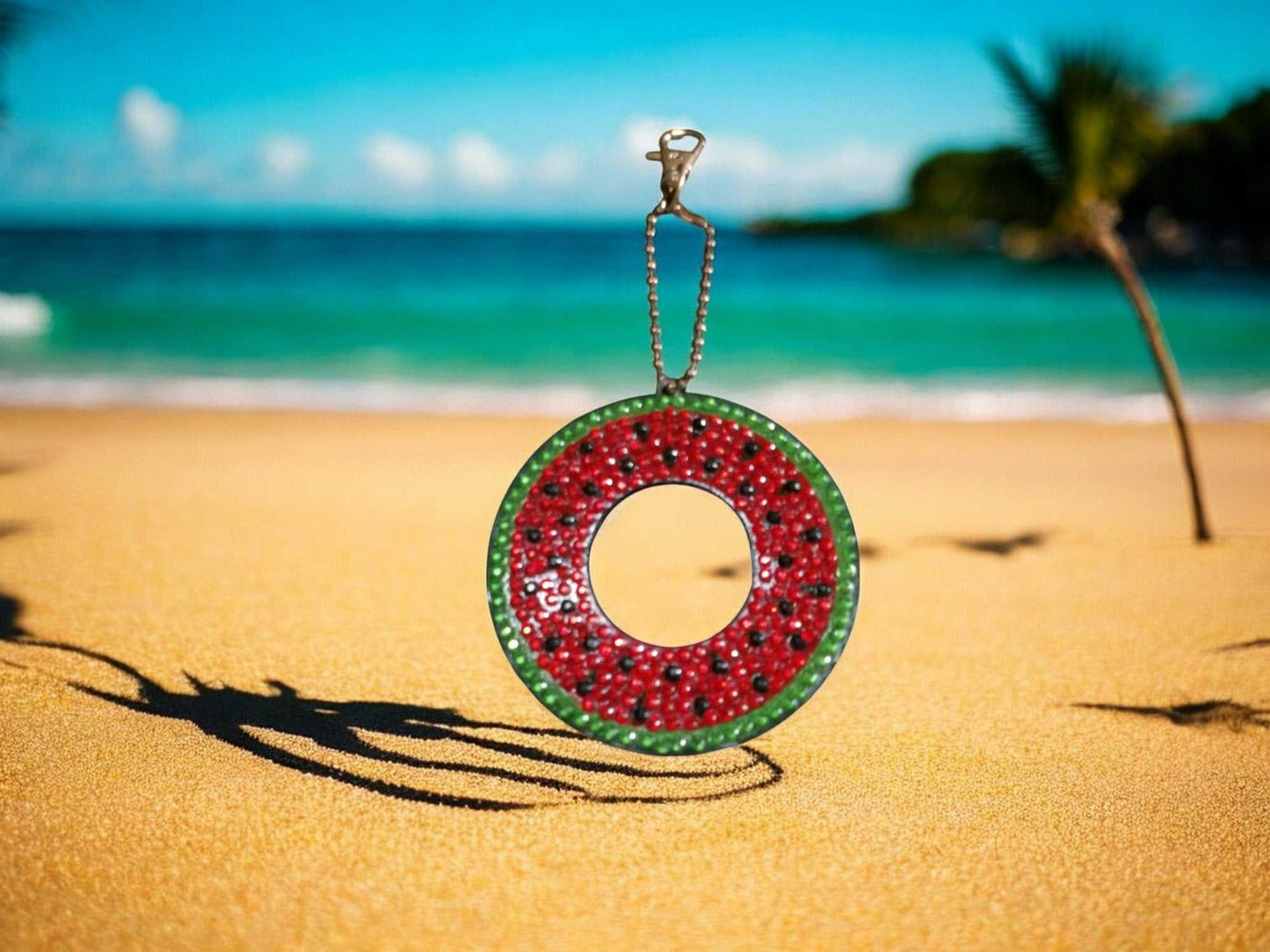 Watermelon keychain with green and red design hanging on beach with ocean background