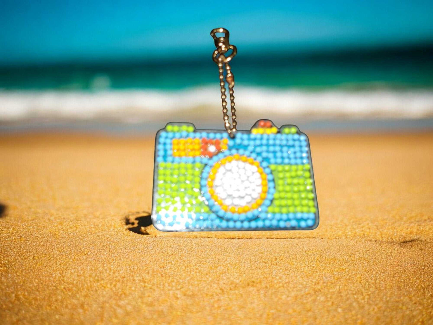 Colorful camera keychain with blue, green, and orange accents on sandy beach background