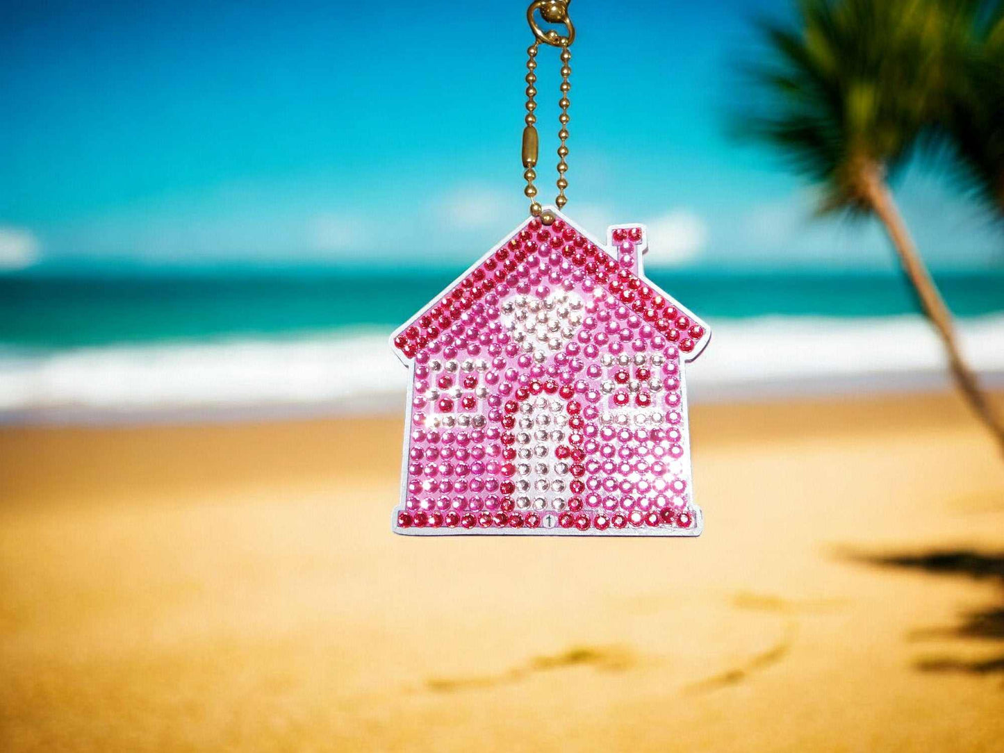 Pink house keychain with heart design hanging on gold chain against beach background