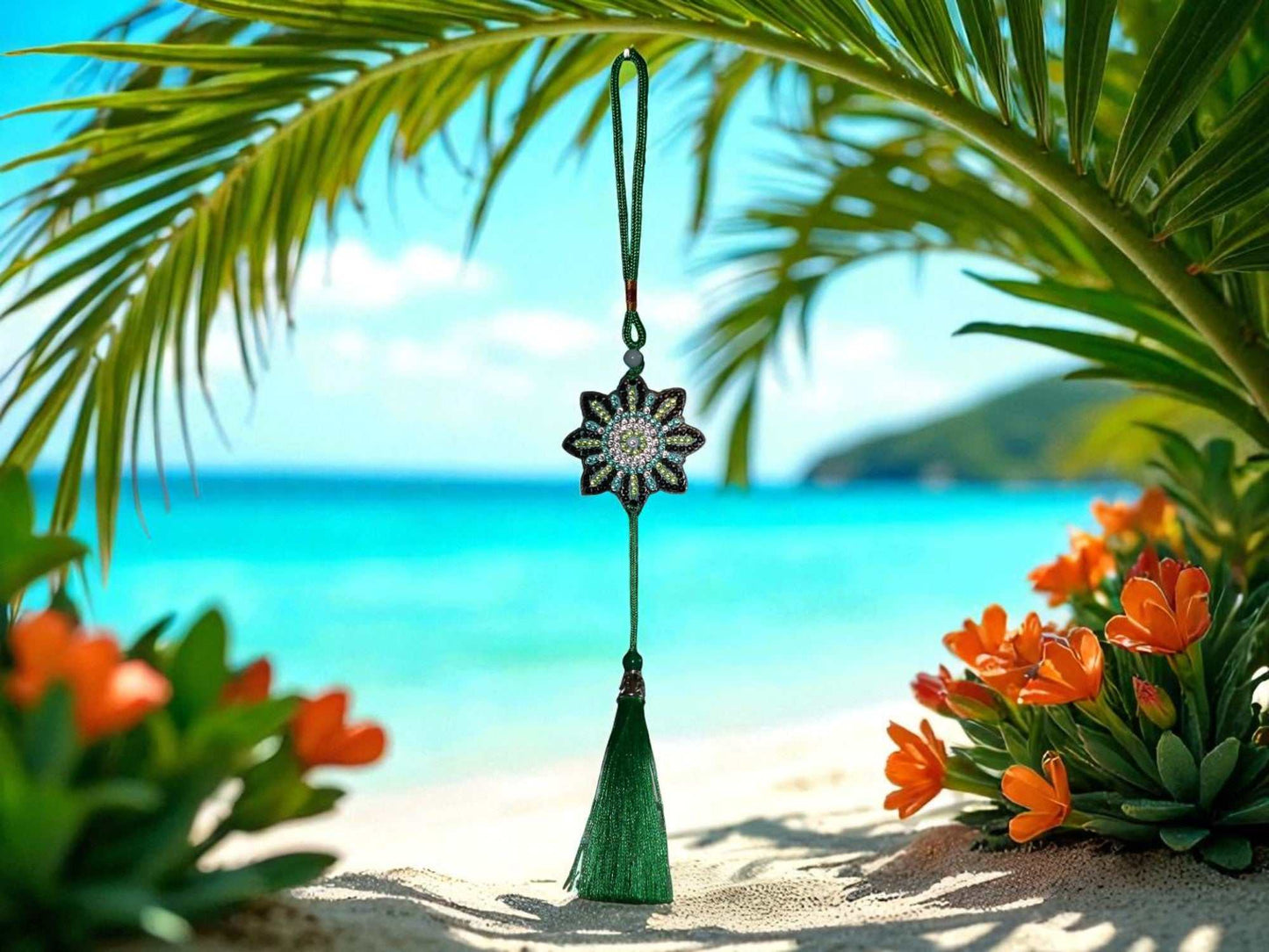 Colorful flower-shaped car ornament with green tassel hanging on beach background with palm leaves and orange flowers.
