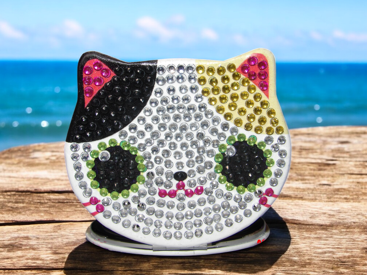 Mixed color cat face pocket mirror with rhinestone details on wooden surface by the sea
