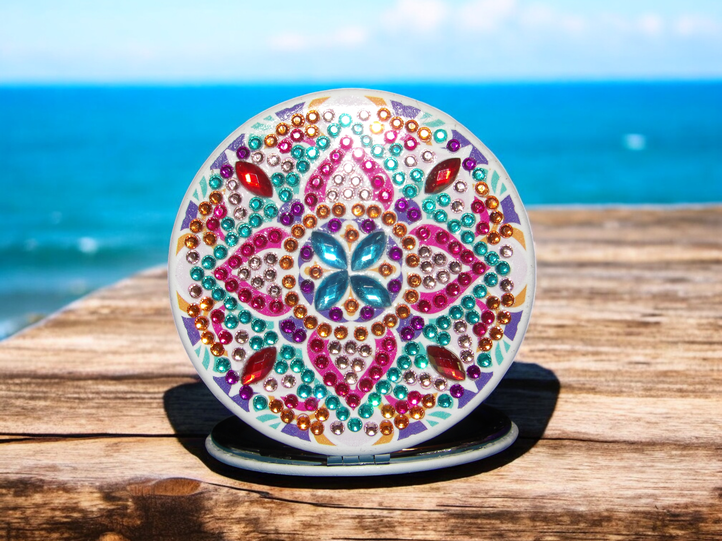Colorful round pocket mirror with mandala design and multicolored gemstones on wooden surface by the sea