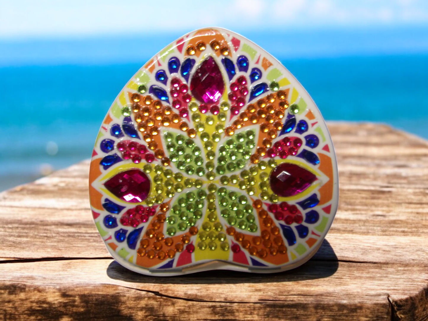 colorful heart shape pocket mirror with mandala design and rhinestones on wooden surface by the sea