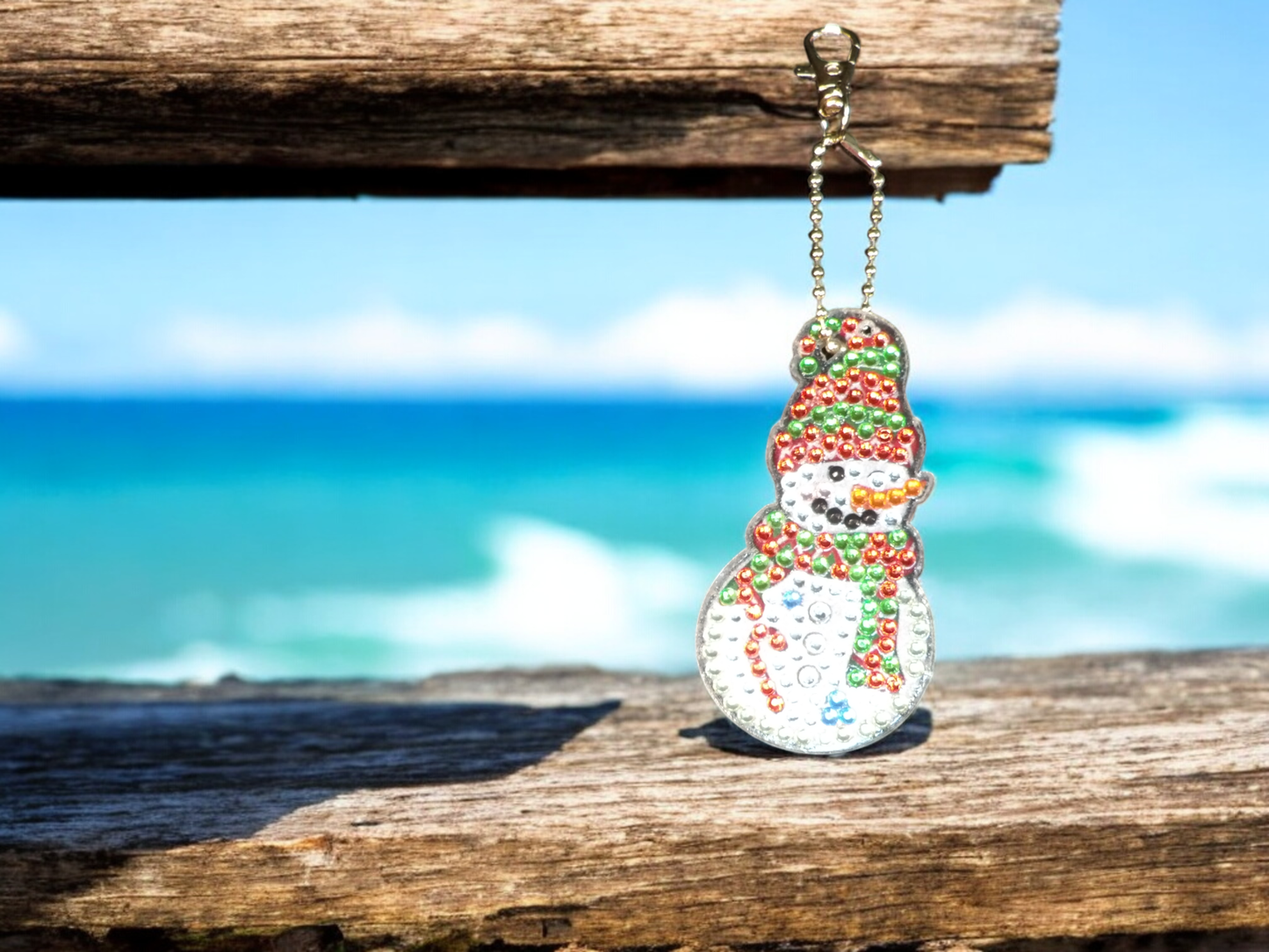 Red and green hat and scarf snowman keychain hanging on wooden surface with blurred ocean background