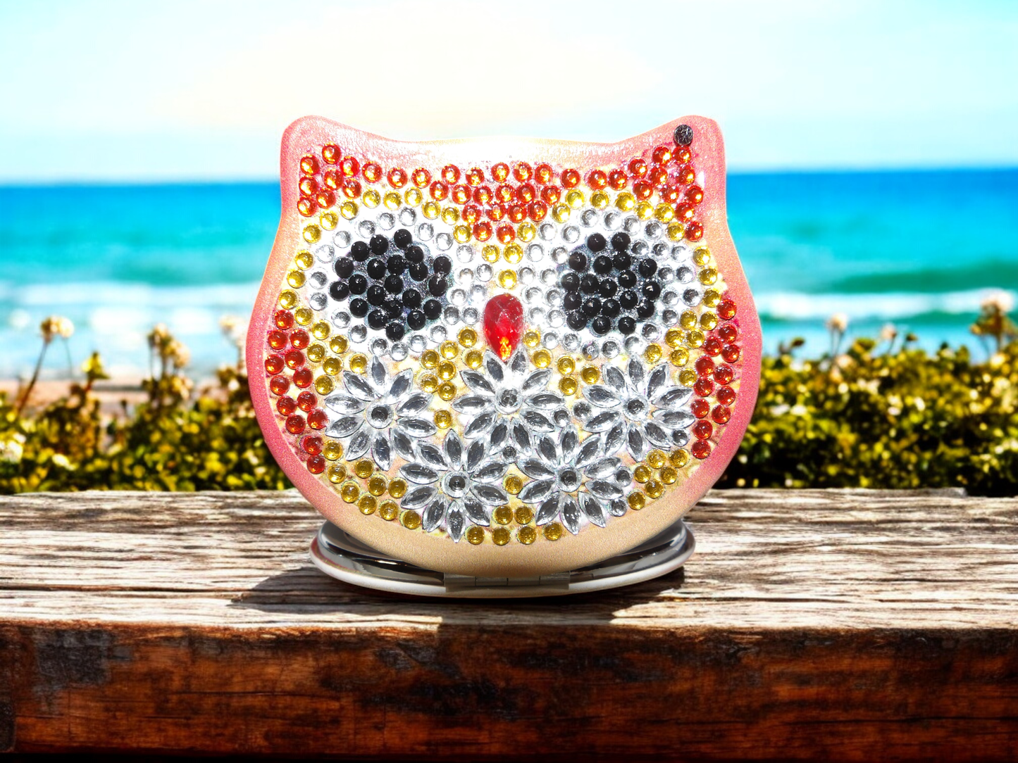 Orange colorful owl pocket mirror with decorative rhinestones and magnification feature placed on wooden surface by the beach