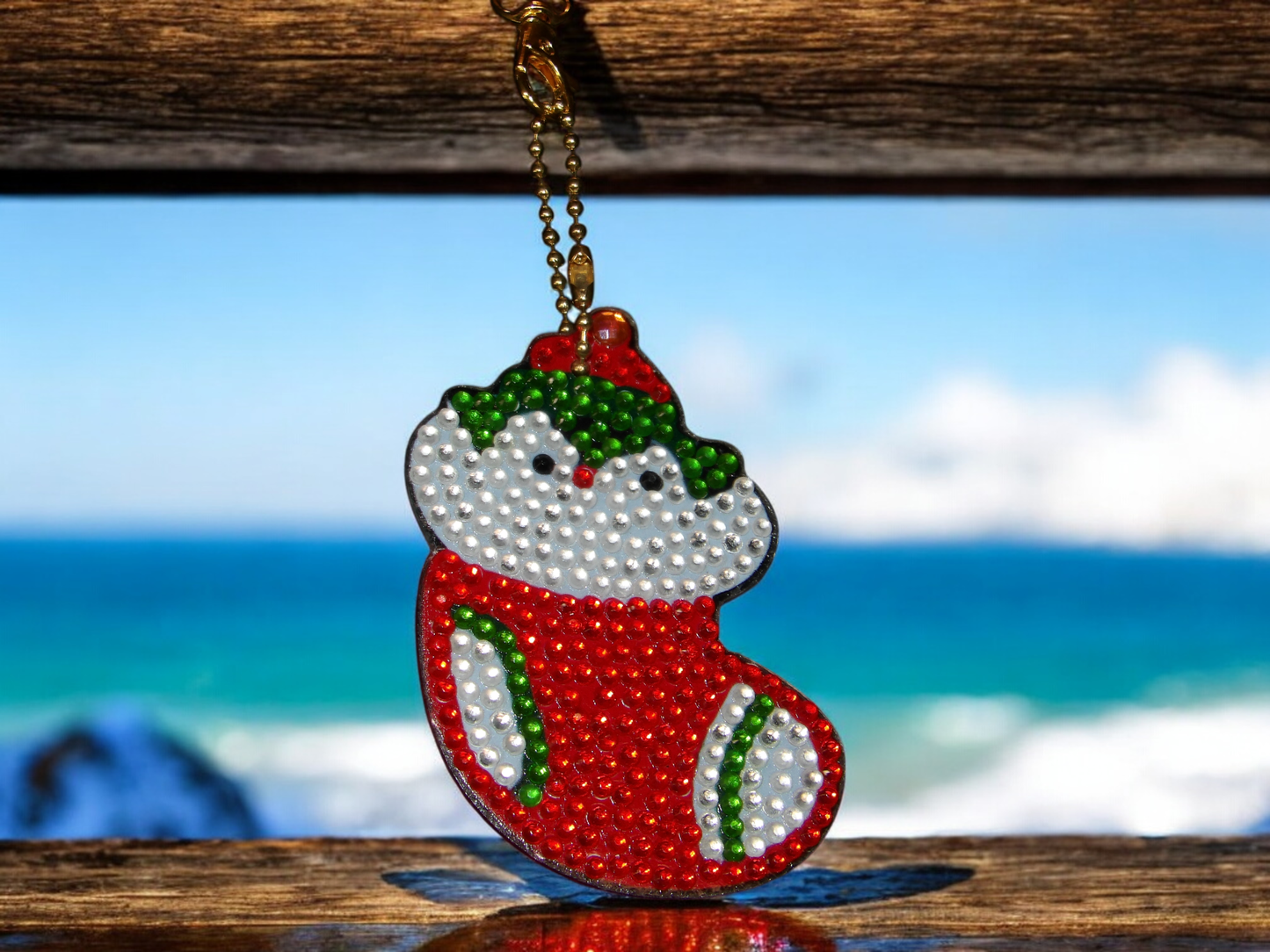 Penguin in Christmas stocking keychain decorated with colorful beads hanging on wooden surface with ocean background