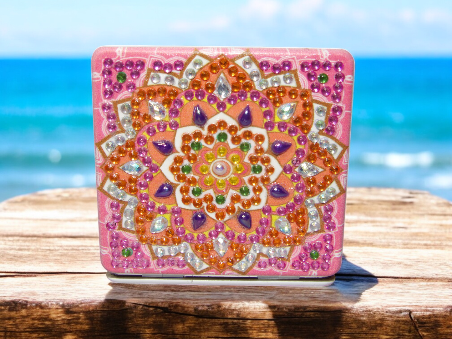 Pink and orange square pocket mirror with mandala design and decorative gemstones on wooden surface by the sea