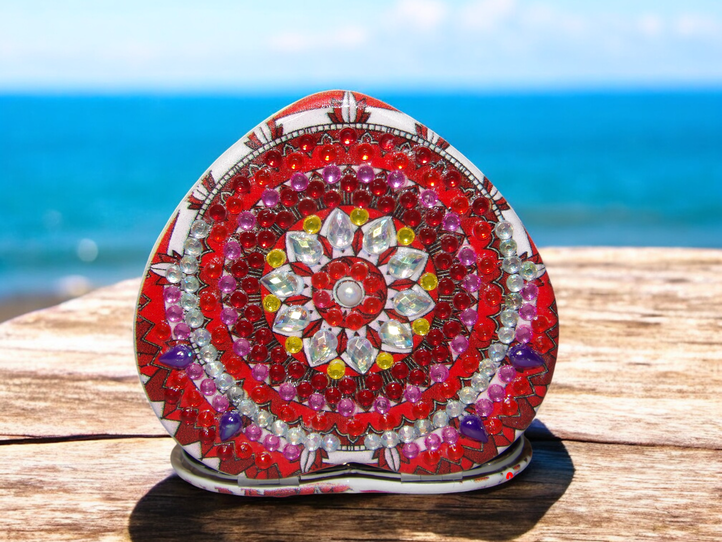 Heart shaped deep red pocket mirror with mandala design and gemstone embellishments on wooden surface by the sea