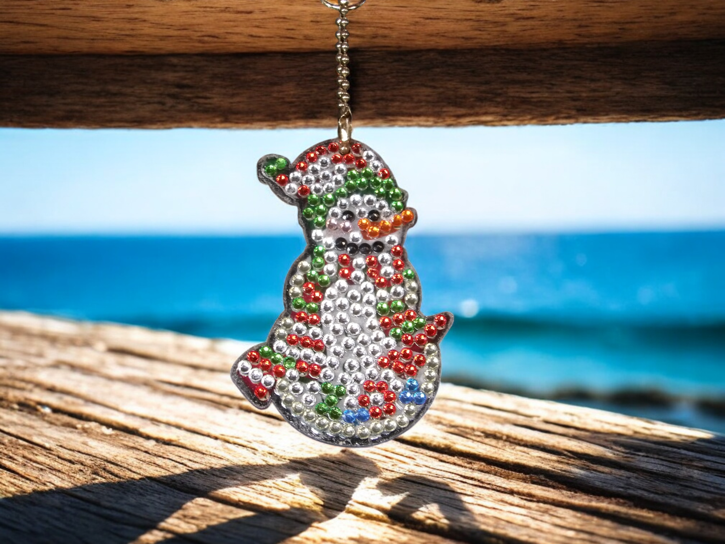 Red and green snowman keychain with beaded design hanging from wooden surface against ocean background