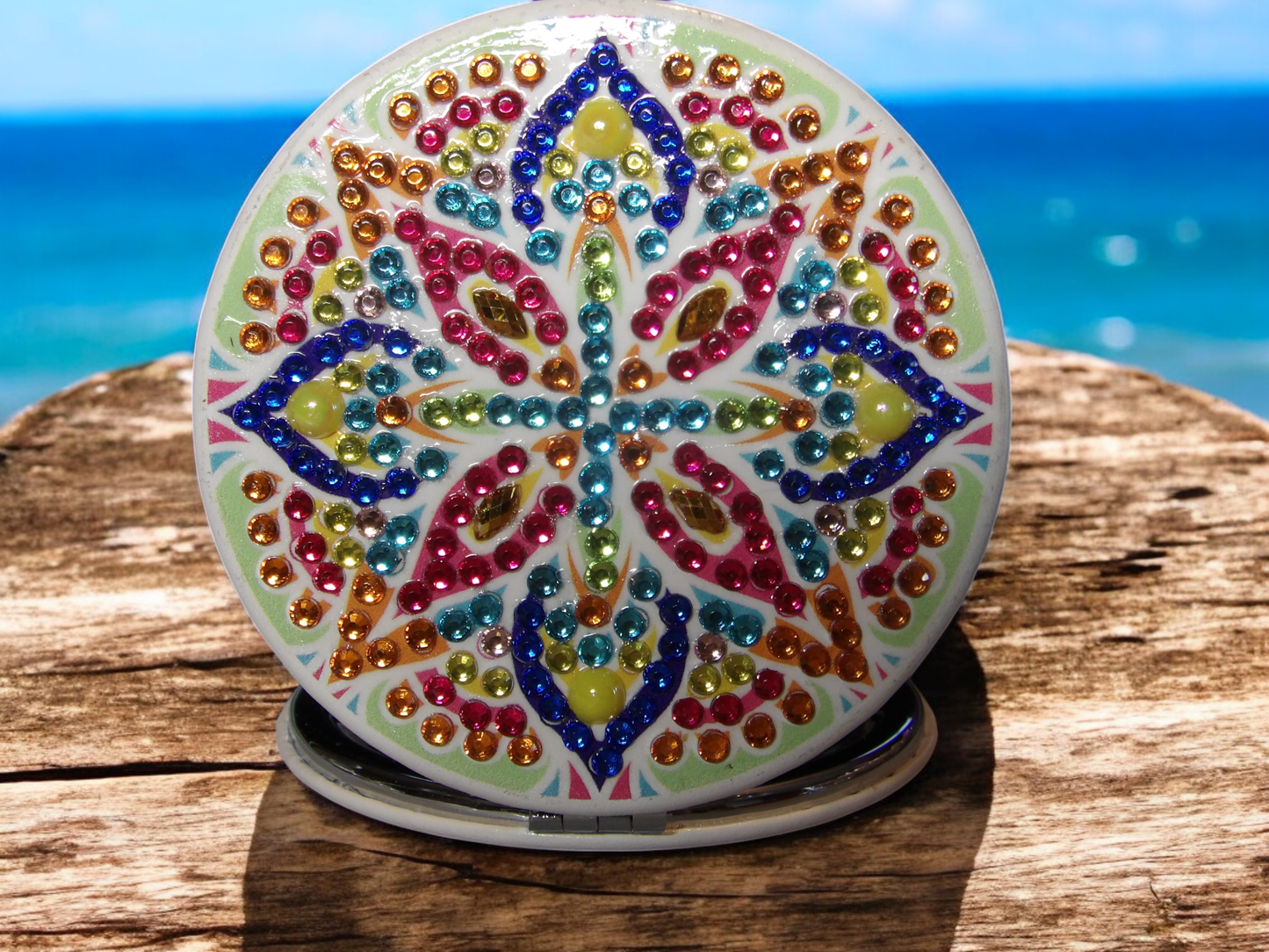 Multi-colored round pocket mirror with mandala design and colorful rhinestones on wooden surface by the sea.